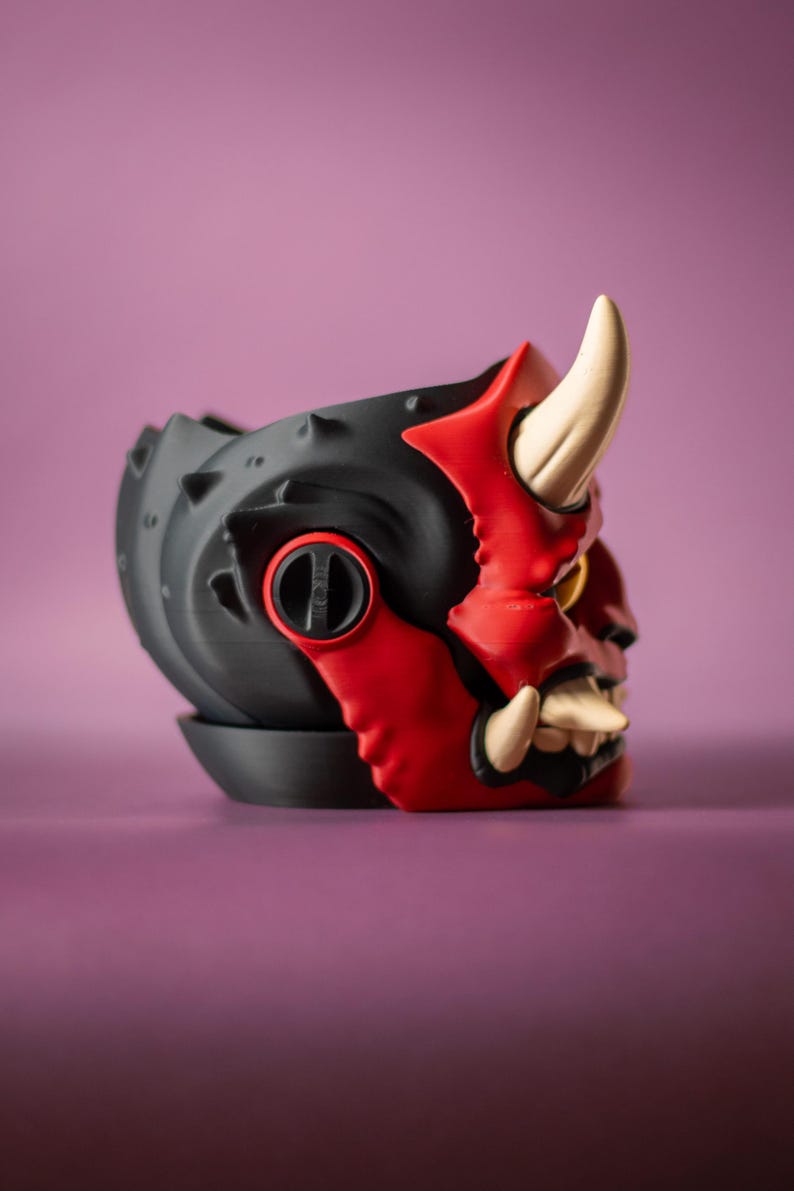 May include: A black and red decorative planter shaped like an Oni mask. The planter features a detailed design with horns, teeth, and a menacing expression. The planter is made of a durable material and is suitable for small plants.