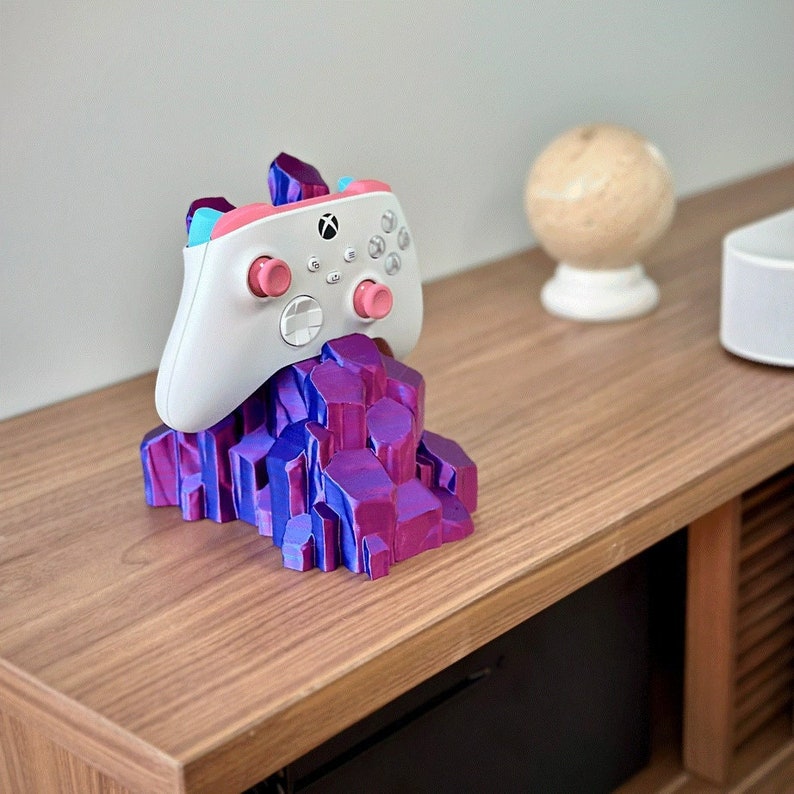 May include: A white video game controller with pink and blue accents sits on a purple and pink crystal-shaped stand. The stand is on a wooden surface.