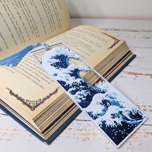 May include: A rectangular bookmark featuring a stylized ocean wave design in shades of blue and white. The bookmark has a blue tassel and is placed in an open book with aged pages. The wave design is a prominent feature.