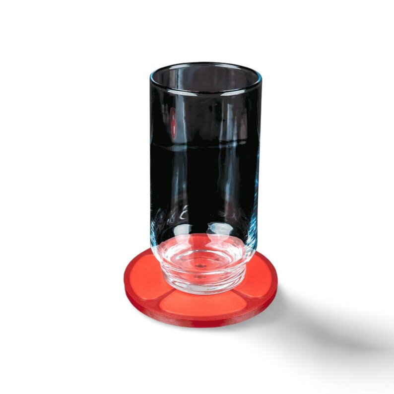 May include: A clear glass tumbler with a dark liquid inside, resting on a bright red, circular coaster. The coaster has a raised, segmented design. The glass is cylindrical with a slightly flared rim, and the coaster is a solid, vibrant red.