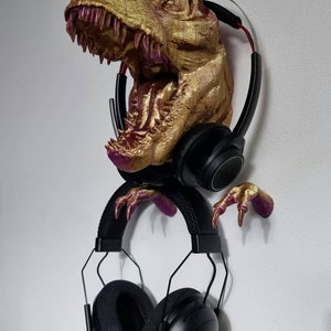May include: A gold-colored dinosaur head with pink accents, mounted on a wall, is used as a headphone holder. Two pairs of black headphones are hanging from the dinosaur's head and claws.