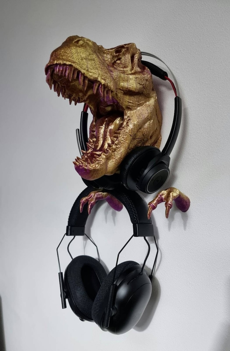 May include: A gold-colored dinosaur head with pink accents, mounted on a wall, is used as a headphone holder. Two pairs of black headphones are hanging from the dinosaur's head and claws.