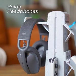 May include: A white headphone stand with a pair of gray headphones hanging on it. The stand also holds a white game controller. The text "Holds Headphones" is visible on the image.