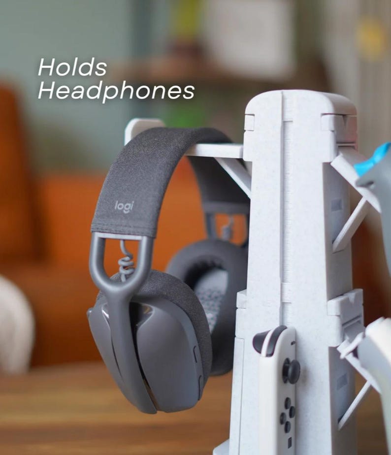 May include: A white headphone stand with a pair of gray headphones hanging on it. The stand also holds a white game controller. The text "Holds Headphones" is visible on the image.
