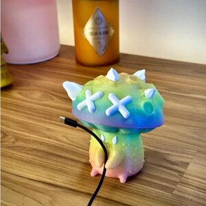 May include: A colorful, rainbow-colored, 3D printed figurine with a mushroom-shaped head and white X's for eyes. The figurine is connected to a black charging cable.