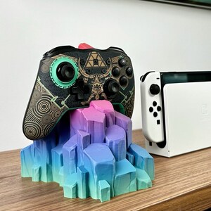 May include: A black and gold video game controller with a green button and a geometric design sits on a colorful, 3D printed stand. A white Nintendo Switch console is in the background.