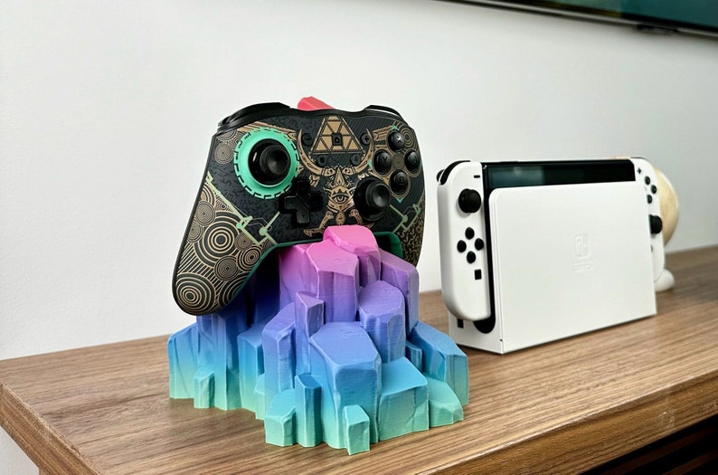 May include: A black and gold video game controller with a green button and a geometric design sits on a colorful, 3D printed stand. A white Nintendo Switch console is in the background.