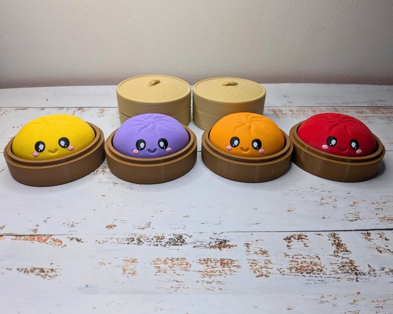 Op de afbeelding: Vier kleurrijke, afgeronde knoedelvormige figuren met lachende gezichten, elk rustend in een bruine ronde basis. De figuren zijn geel, paars, oranje en rood. Twee beige, ronde deksels zijn achter de figuren geplaatst.