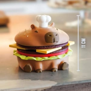 Op de afbeelding: Een grillige, bruine capibara-vormige container ontworpen als een hamburger, met een koksmuts. De hamburger heeft lagen kaas, een rode plak tomaat en sla. Het meet 11 cm in hoogte.