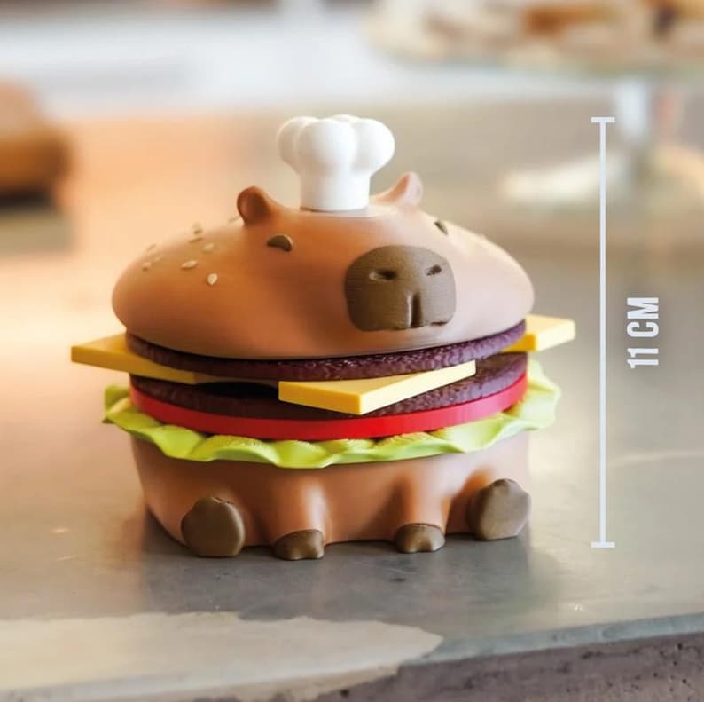 Op de afbeelding: Een grillige, bruine capibara-vormige container ontworpen als een hamburger, met een koksmuts. De hamburger heeft lagen kaas, een rode plak tomaat en sla. Het meet 11 cm in hoogte.