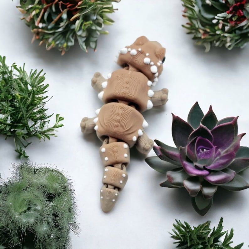 May include: A brown and white segmented toy lizard with a textured surface. The toy is posed on a white background with green plants.
