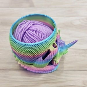 May include: A rainbow-colored unicorn yarn bowl with a purple ball of yarn inside. The bowl is made of 3D printed plastic and has a textured surface.