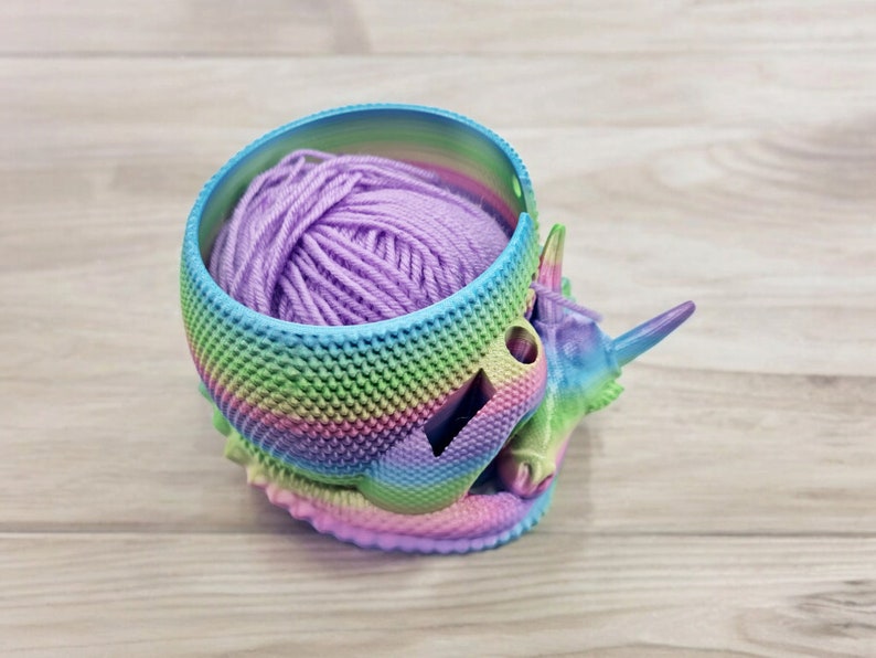 May include: A rainbow-colored unicorn yarn bowl with a purple ball of yarn inside. The bowl is made of 3D printed plastic and has a textured surface.