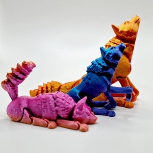 May include: Three 3D printed wolf figurines in different poses. The wolf on the left is pink, the middle wolf is blue, and the wolf on the right is orange.