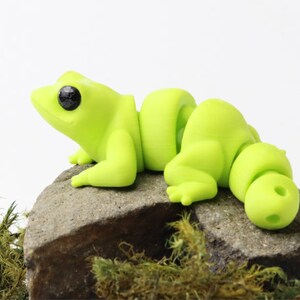 May include: A lime green segmented frog figurine with black eyes, sitting on a gray rock with green moss.