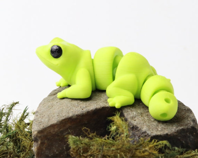 May include: A lime green segmented frog figurine with black eyes, sitting on a gray rock with green moss.