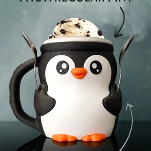 May include: A black, white, and orange penguin-shaped mug with two spoons. The mug is filled with ice cream. Text on the image reads "FITS A REGULAR PINT AND 2 SPOONS."