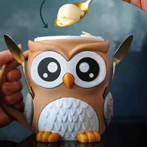 May include: An owl-shaped ice cream bowl with a lid, holding two gold spoons. The bowl is brown, white, and orange, with large black and white eyes. Text on the image reads "FITS A REGULAR PINT" and "AND HOLDS TWO SPOONS".