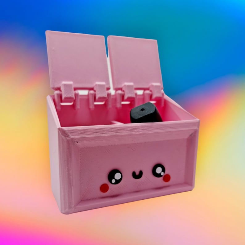 May include: A pink plastic box with a hinged lid and a cute cartoon face. The box has a small black compartment inside.