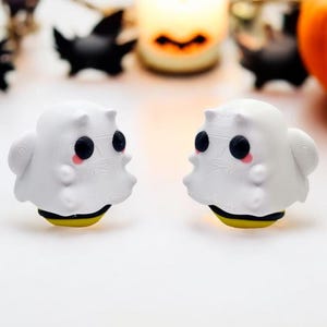 May include: Two white ghost-shaped Halloween ornaments with black eyes and pink cheeks. Each ghost is wearing a yellow and black striped headband.