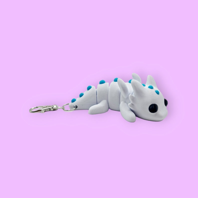 May include: White dragon keychain with blue accents. The dragon is lying on its belly with its wings spread out. It has a silver key ring attached to its tail.