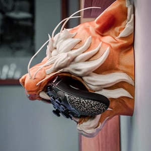 May include: A wall-mounted tiger head sculpture in shades of orange and white, holding a black and gray patterned game controller in its mouth. The sculpture is mounted on a wooden surface.