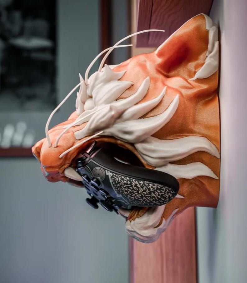 May include: A wall-mounted tiger head sculpture in shades of orange and white, holding a black and gray patterned game controller in its mouth. The sculpture is mounted on a wooden surface.