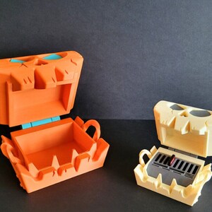 May include: Two open skull-shaped boxes. The box on the left is orange with a teal hinge. The box on the right is cream colored with a black interior.