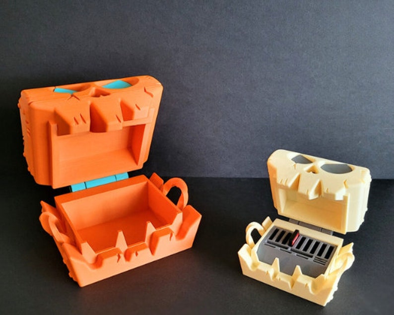 May include: Two open skull-shaped boxes. The box on the left is orange with a teal hinge. The box on the right is cream colored with a black interior.