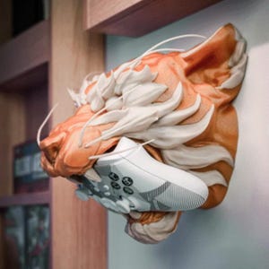 Puede incluir: Escultura de una cabeza de tigre naranja y blanca sosteniendo un mando de videojuegos blanco y gris. La escultura está montada en una pared. El mando tiene un agarre texturizado. La cabeza del tigre tiene detalles.