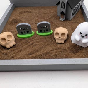 Pu&ograve; includere: Una sandbox grigia con una piccola chiesa grigia, due lapidi grigie con il testo "RIP", due teschi marroni e un fantasma bianco con un viso sorridente.