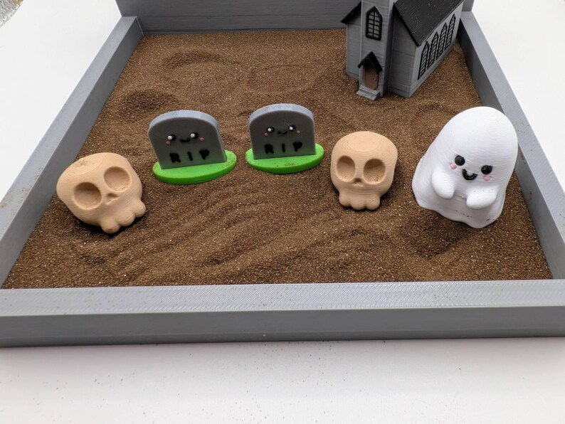 Pu&ograve; includere: Una sandbox grigia con una piccola chiesa grigia, due lapidi grigie con il testo "RIP", due teschi marroni e un fantasma bianco con un viso sorridente.