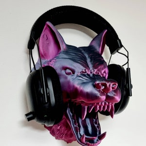 May include: A 3D-printed wolf head in shades of purple and gray, with an open mouth revealing sharp teeth. Black headphones are placed over the head. The wolf head is mounted on a white wall.