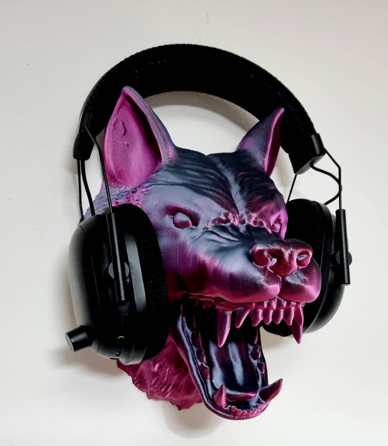 May include: A 3D-printed wolf head in shades of purple and gray, with an open mouth revealing sharp teeth. Black headphones are placed over the head. The wolf head is mounted on a white wall.