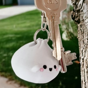 May include: A white, round, plush keychain with a cute cartoon face. The keychain is attached to a silver key ring and a silver key.