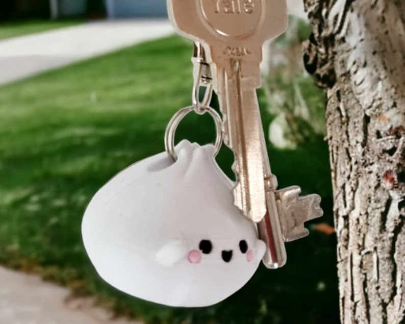 May include: A white, round, plush keychain with a cute cartoon face. The keychain is attached to a silver key ring and a silver key.