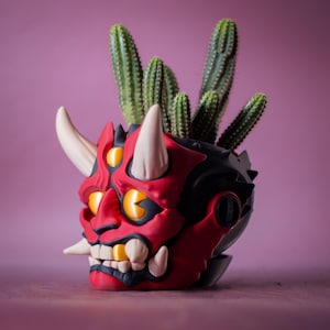 May include: A red and black decorative planter shaped like a demon's head, with white horns and teeth, and yellow eyes. The planter holds several green cacti. The background is a solid pink color.