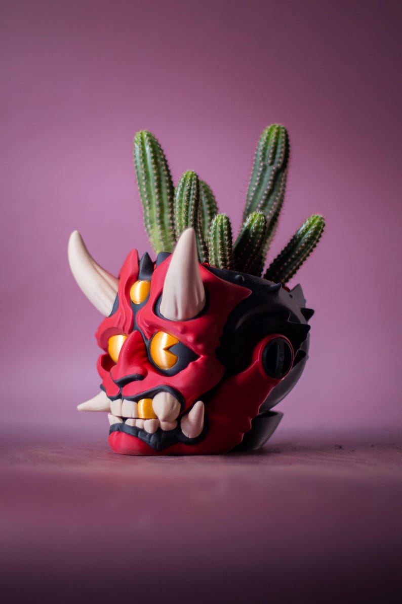 May include: A red and black decorative planter shaped like a demon's head, with white horns and teeth, and yellow eyes. The planter holds several green cacti. The background is a solid pink color.