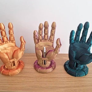 May include: Three 3D printed robot hand sculptures, each with a different color finish. The hands are posed with fingers extended and are mounted on a circular base. The colors are gold, purple, and teal.