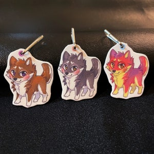May include: Three keychains featuring cartoon wolf designs. The wolf on the left is brown with white fur, the middle wolf is gray with white fur, and the wolf on the right is red, yellow, and purple.