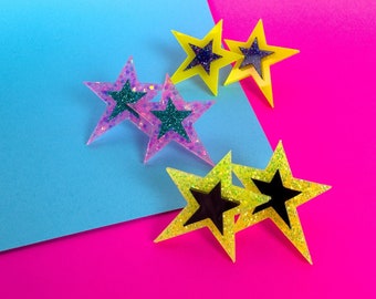 Può includere: Cinque orecchini a forma di stella con glitter in vari colori. Due stelle gialle con centri neri, due stelle viola con centri turchesi e una stella turchese con un centro viola.