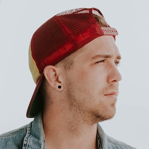 May include: A man wearing a red and yellow baseball cap with a black earring. He is looking to the right and has a beard.