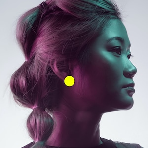 May include: A woman with a purple and blue hairstyle is wearing a bright yellow circular earring. The earring is in her left ear.