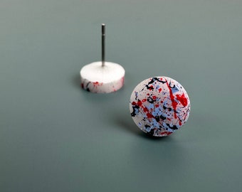 May include: A pair of white stud earrings with a red, blue and black splatter paint design.