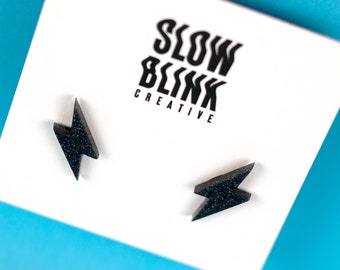 May include: A pair of black glitter lightning bolt stud earrings. The earrings are on a white card with the text "Slow Blink Creative".