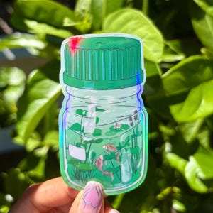 May include: A clear glass jar with a green lid, filled with water, plants, and two small orange fish. The jar is a sticker with a holographic finish.