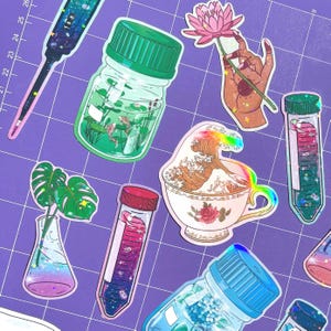 May include: A set of colourful stickers featuring various designs, including a glass jar with fish, a test tube with a galaxy inside, a teacup with a wave, a hand holding a flower, and a monstera plant in a beaker. The stickers are on a purple grid background.