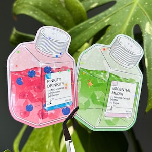 May include: Two stickers shaped like bottles, one pink and one green, with white screw-top lids. The pink bottle says "PINKITY DRINKITY" and contains blueberries. The green bottle says "ESSENTIAL MEDIA". Both have ingredient lists.