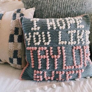 May include: A gray crocheted pillow with the text "I know you think you're so tough" in white and pink yarn. The pillow is sitting on a white bed with other pillows.