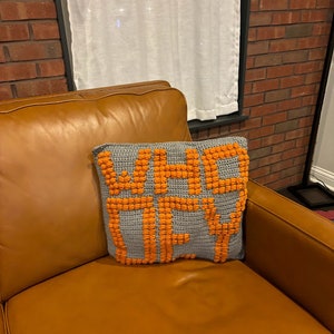 May include: A gray crochet pillow with the words "WHO FEY" in orange yarn. The pillow is rectangular and has a textured, raised design. It rests on a brown leather couch.